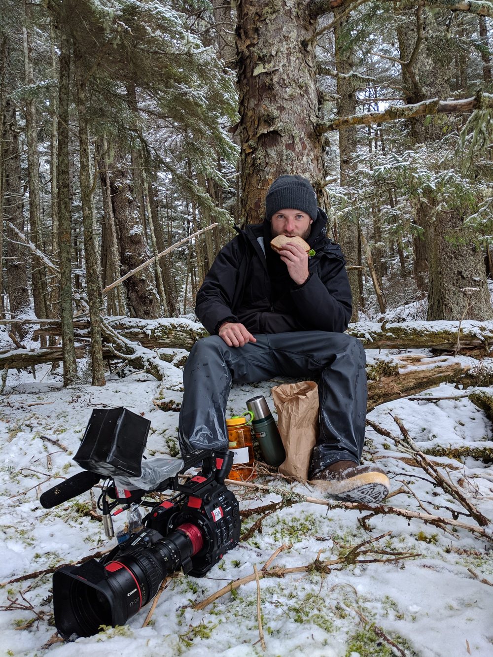 Dwayne Fowler eating a sandwich on a snowy log beside his camera